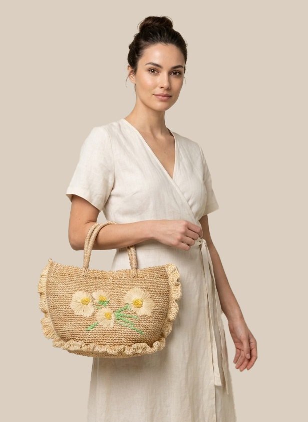 A woman wearing a cream linen wrap dress holds a natural straw tote bag decorated with embroidered yellow flowers and green leaves, set against a solid beige background.