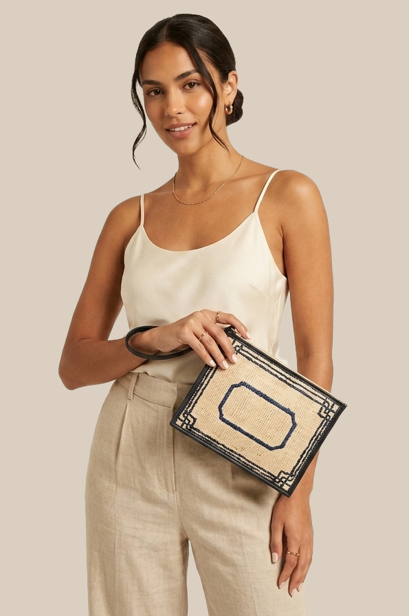 A woman wearing a cream camisole and beige linen trousers holds a woven clutch bag with a black border and blue geometric pattern against a solid beige background.
