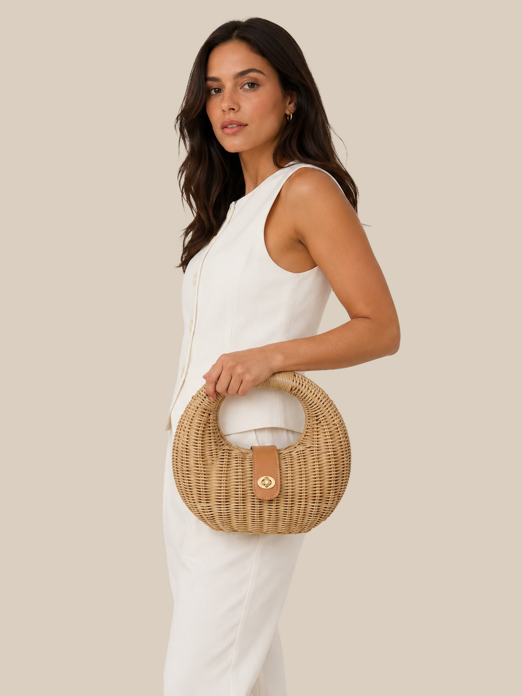 A woman in a white sleeveless jumpsuit holds a round woven straw handbag with a tan leather handle and front strap against a solid beige background.