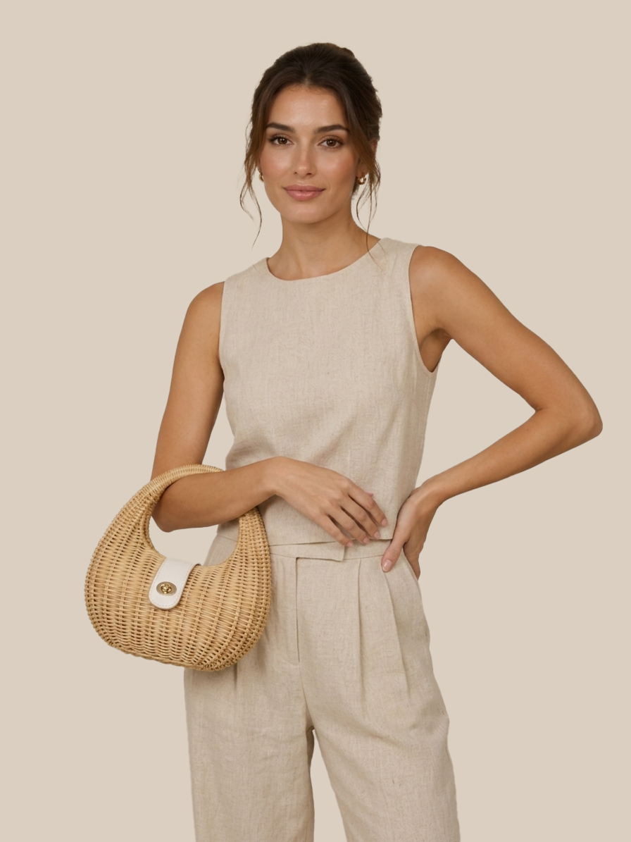 A woman modeling a sleeveless beige linen jumpsuit with matching trousers, holding a woven straw crescent handbag with a white button clasp against a solid beige background.