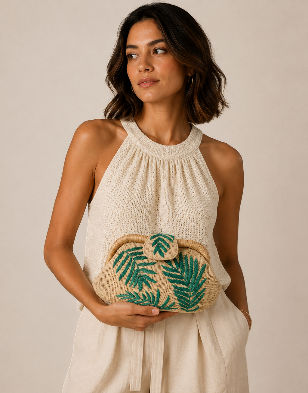 A woman wearing a cream halter top holds a woven straw clutch bag decorated with green palm leaf embroidery, set against a plain beige background.