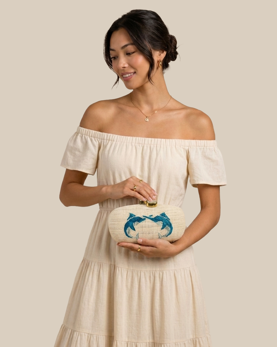 A woman wearing an off-the-shoulder cream dress holds a cream clutch purse decorated with a blue dragon design and gold hardware, set against a solid beige background.