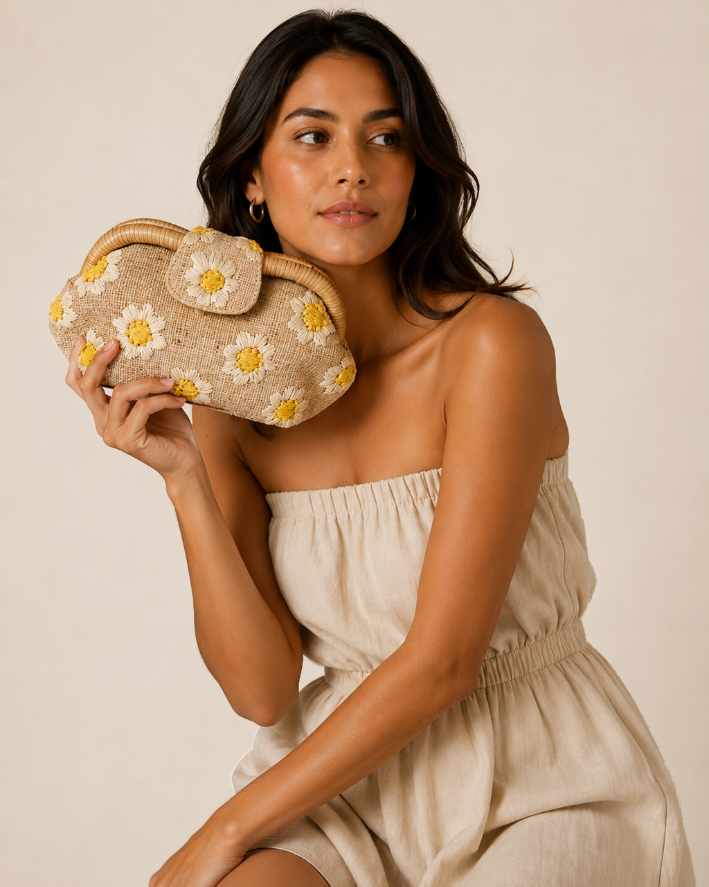 A woman in a beige strapless dress holds a woven clutch bag decorated with white and yellow daisy flowers against a plain beige background.