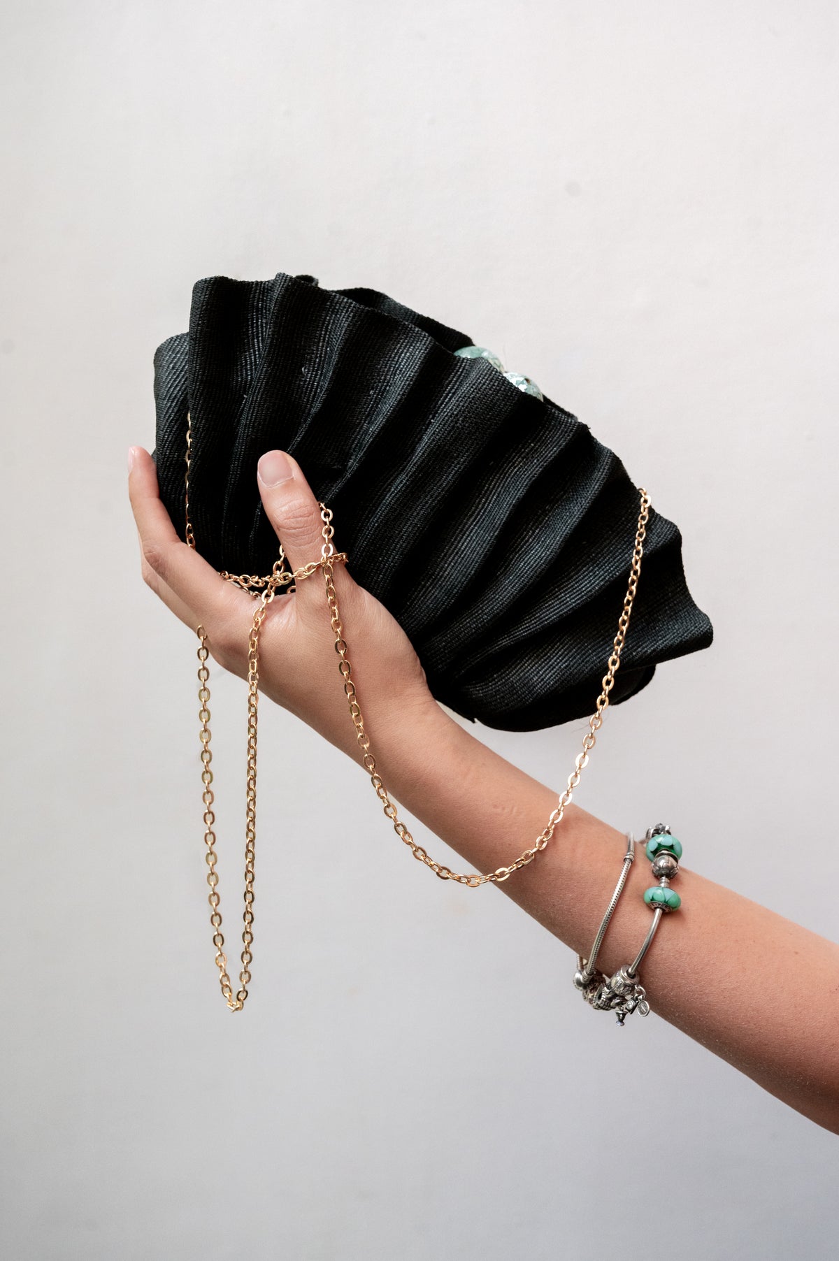 Handheld view of the Josephine Clamshell Clutch Black held in hand with its gold-tone chain strap draped gracefully against a neutral wall.